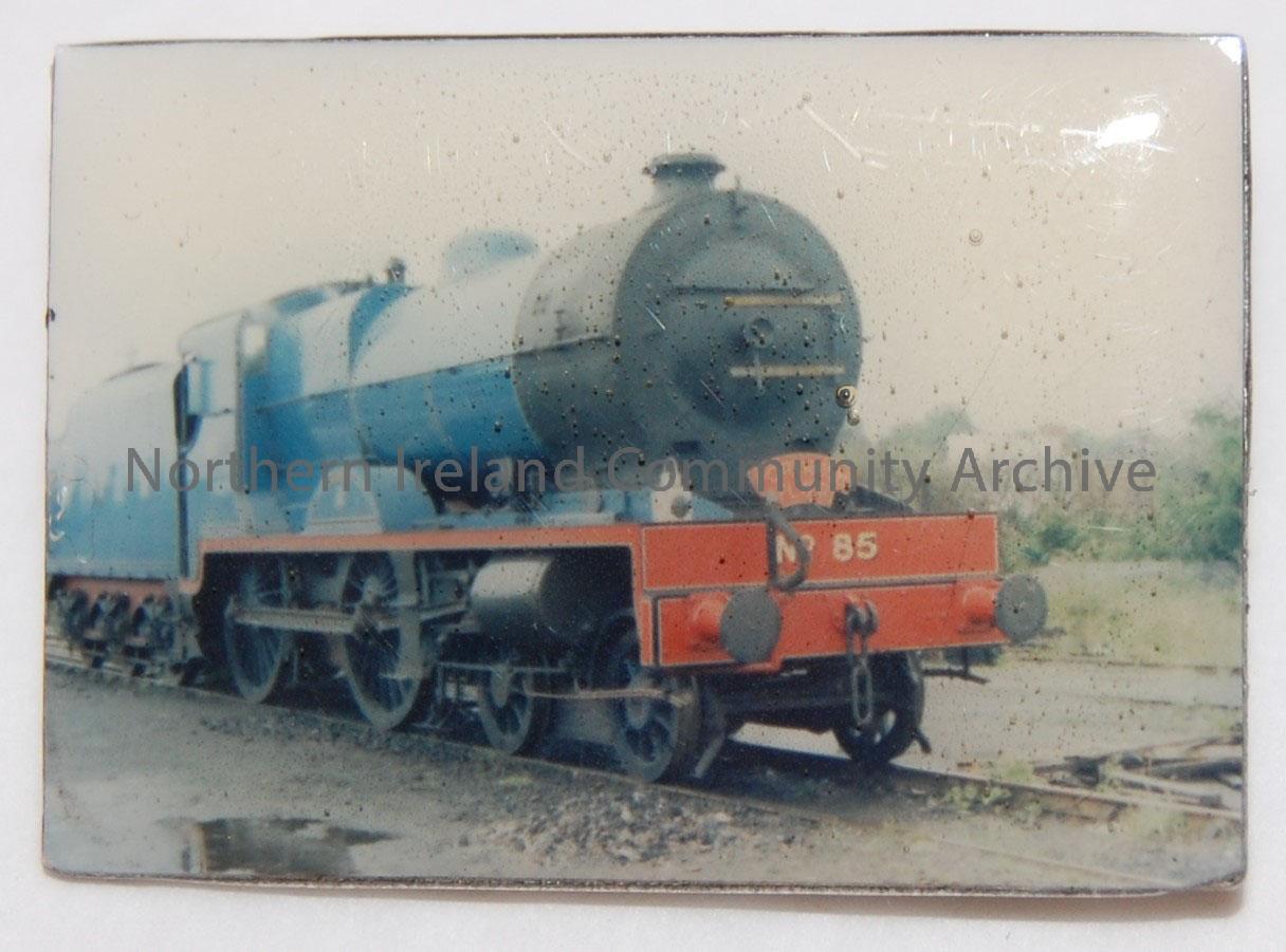 Railway badge commemorating the 150th Anniversary of the Ulster Railway. Colour photograph of a blue engine. In plastic box, with label on bottom – 2007.16 (1)