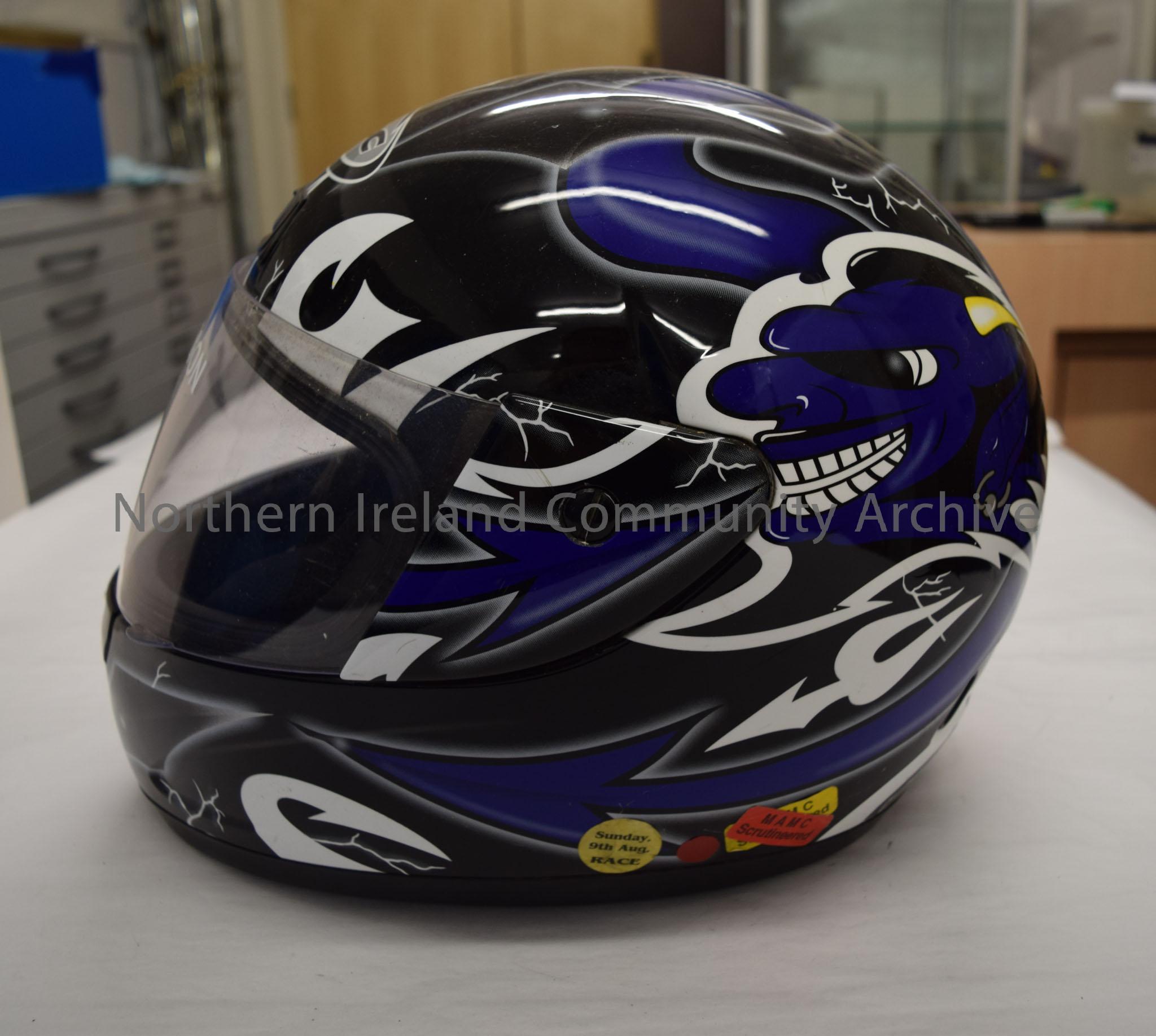 KBC motorcycle helmet belonging to Lee Jackson. Black with dark blue and white pattern.Image of a goblin’s head with yellow and white horn and arrow p… – 2016.55 (3)