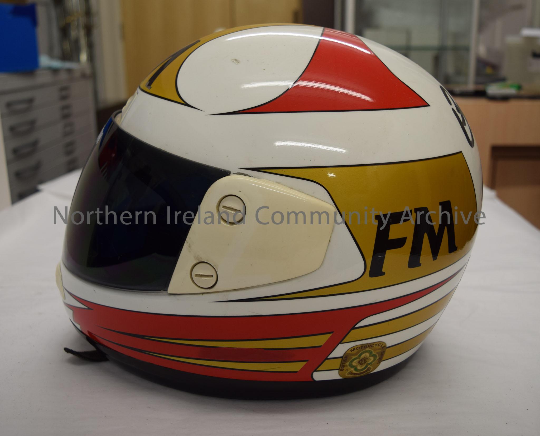 FM motorcycle helmet belonging to Brian Morrison. White helmet with gold and red pattern and gold and red stripes along the bottom. FM written in blac… – 2016.58 (3)