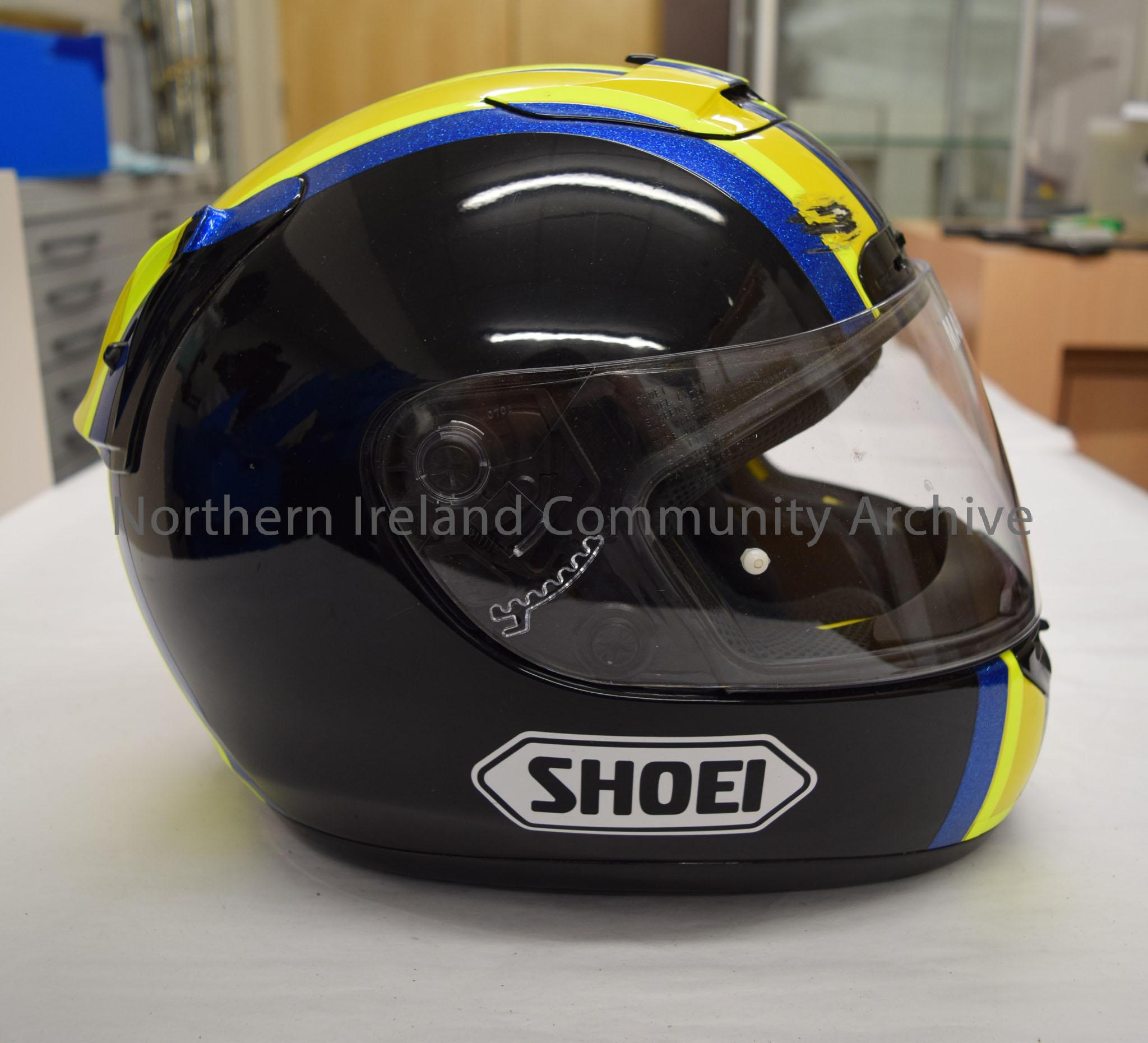 Shoei motorcycle helmet belonging to Marty Nutt. Black helmet with a yellow, fluorescent yellow and sparkly blue “M” pattern on the top.  – 2016.84 (5)