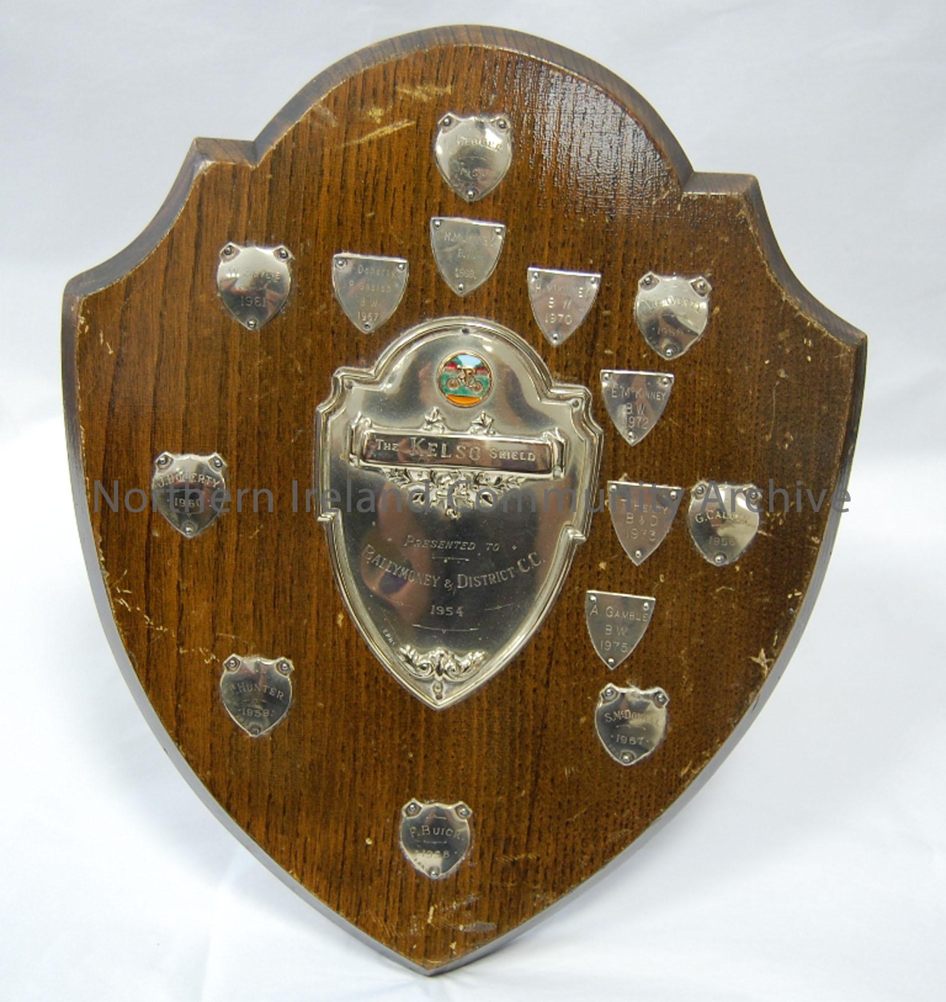 Wooden shield with smaller metal plaques naming winners of shield 1954-1975. Central shield engraved ‘The Kelso shield presented to Ballymoney & Distr…