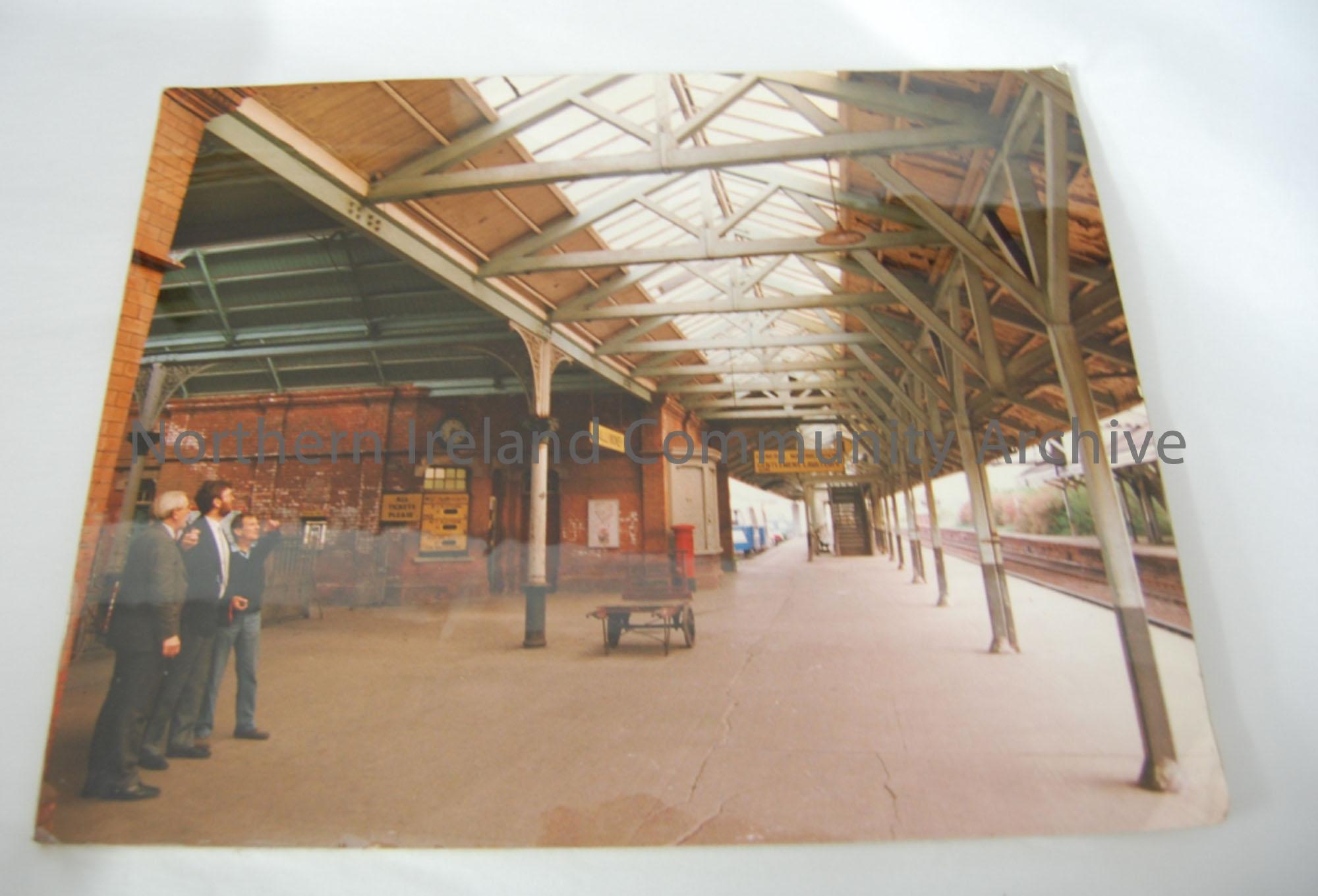 photograph on spongeboard of the platform of Ballymoney train station- three men are standing on the platform and there is a cart on the platform