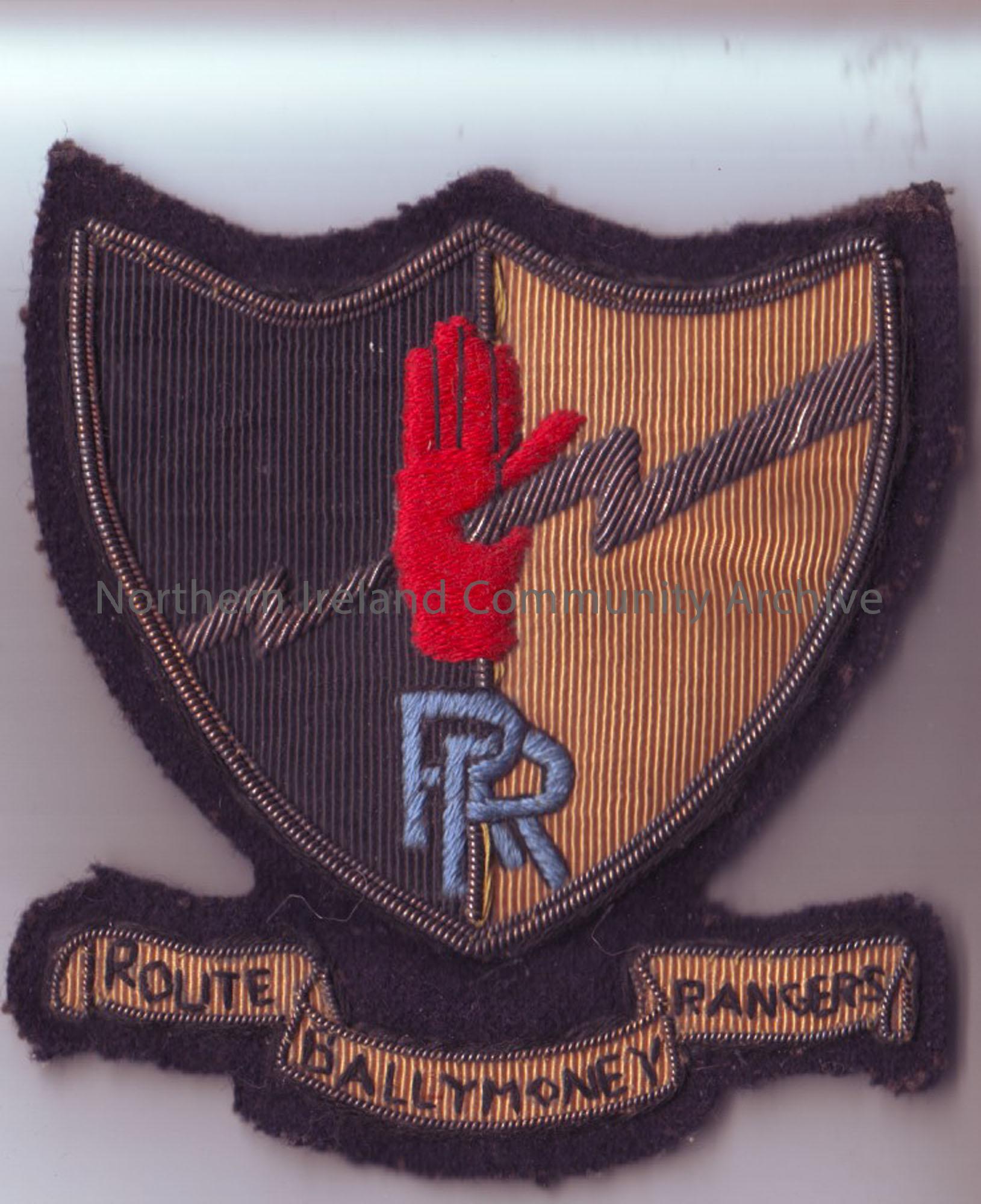 Embroidered badge of “Route Ballymoney Rangers.” Badge is a shield shape with a half black and half yellow background. In the middle of the shield is …