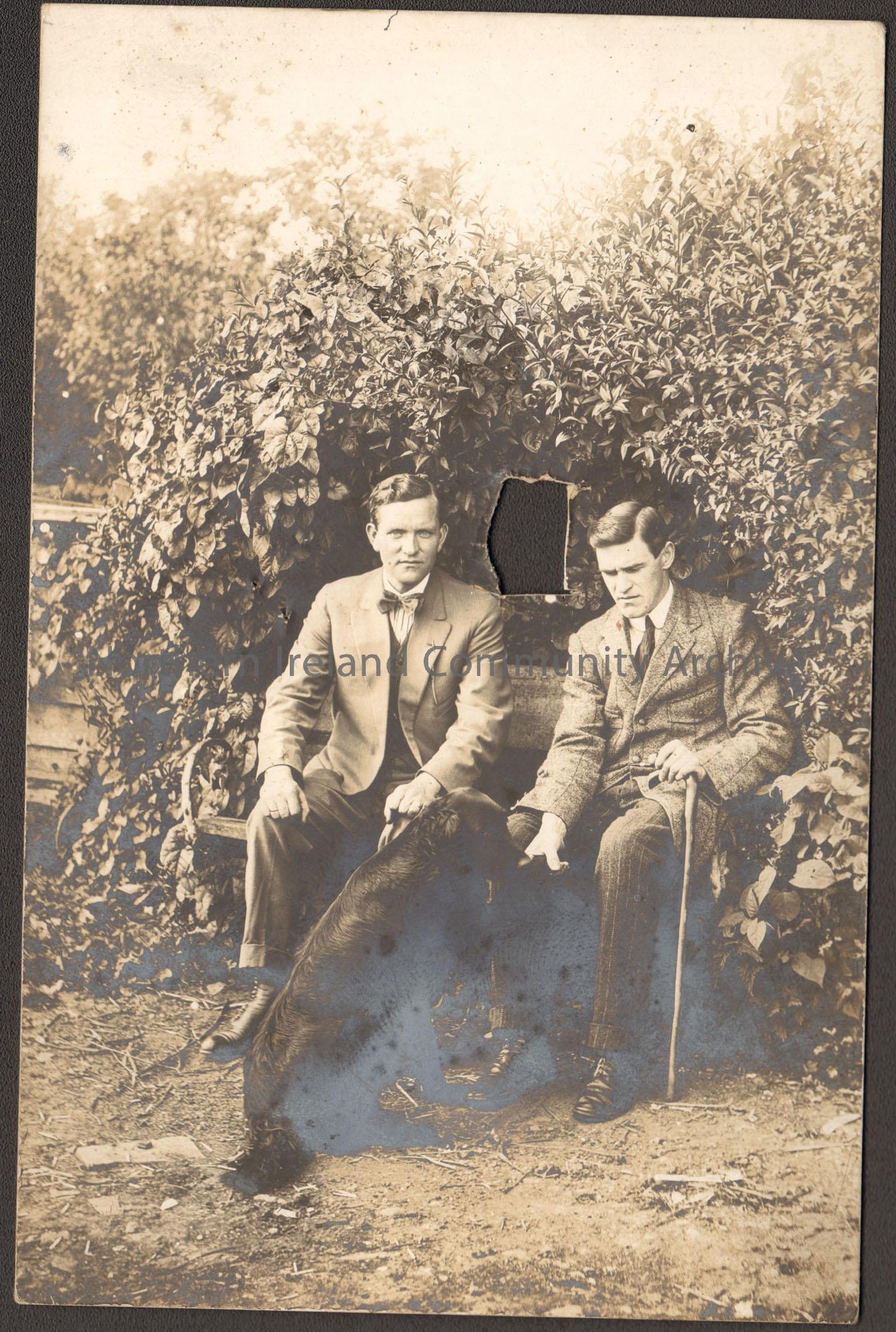Black and white school photograph of two men sitting in a garden with a dog. On right, George Shiels.