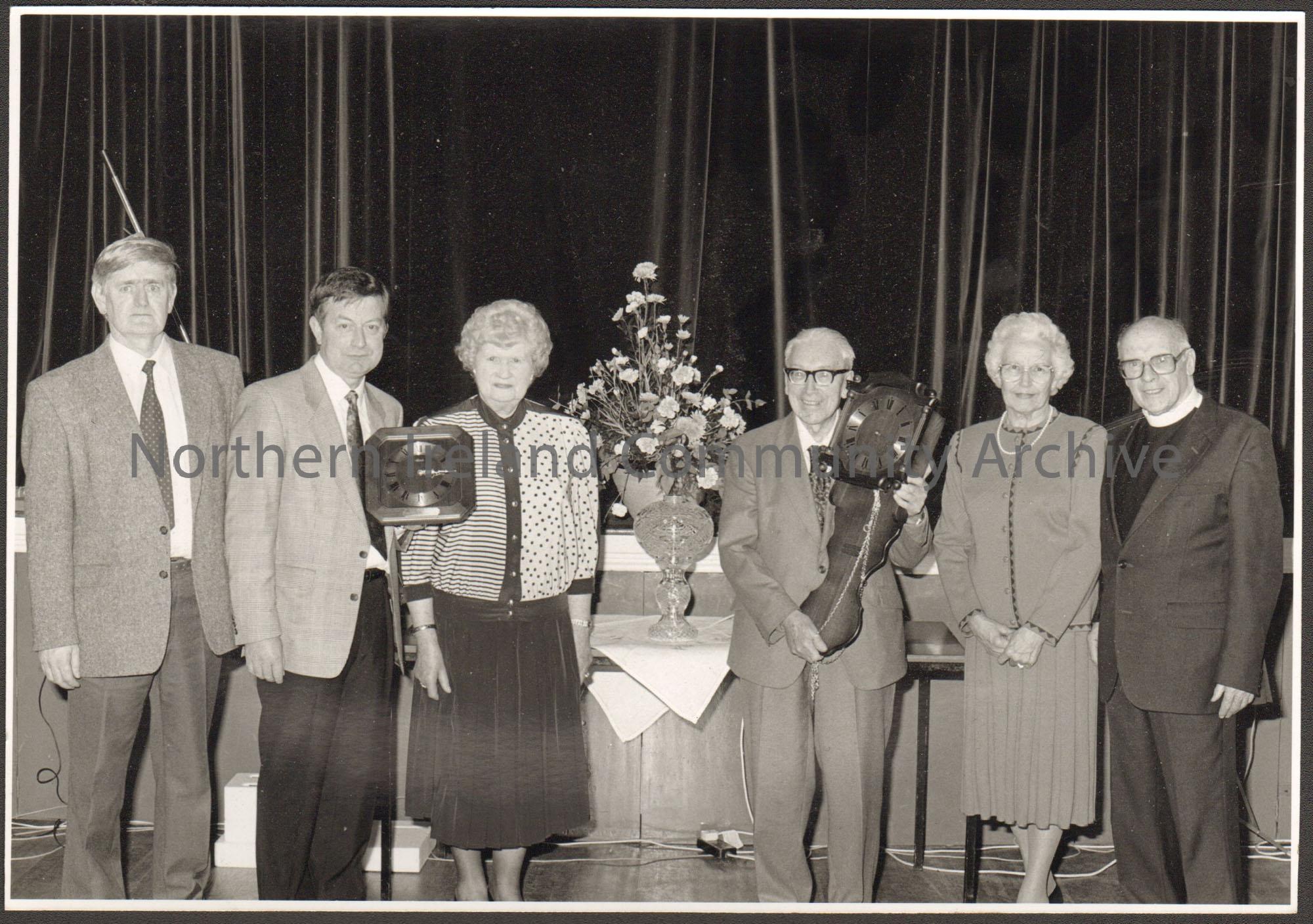 Black and white photograph of six people, including Molly Holmes and James McAfee. Two people are holding clocks. Taken in the Town hall auditorium