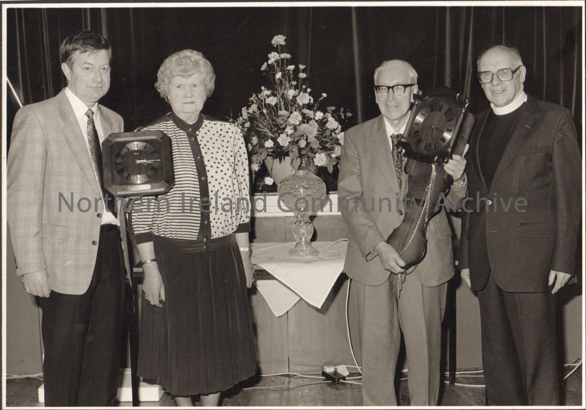 Black and white photograph of four people, including Molly Holmes and James McAfee. Two people are holding clocks. Taken in the Town hall auditorium