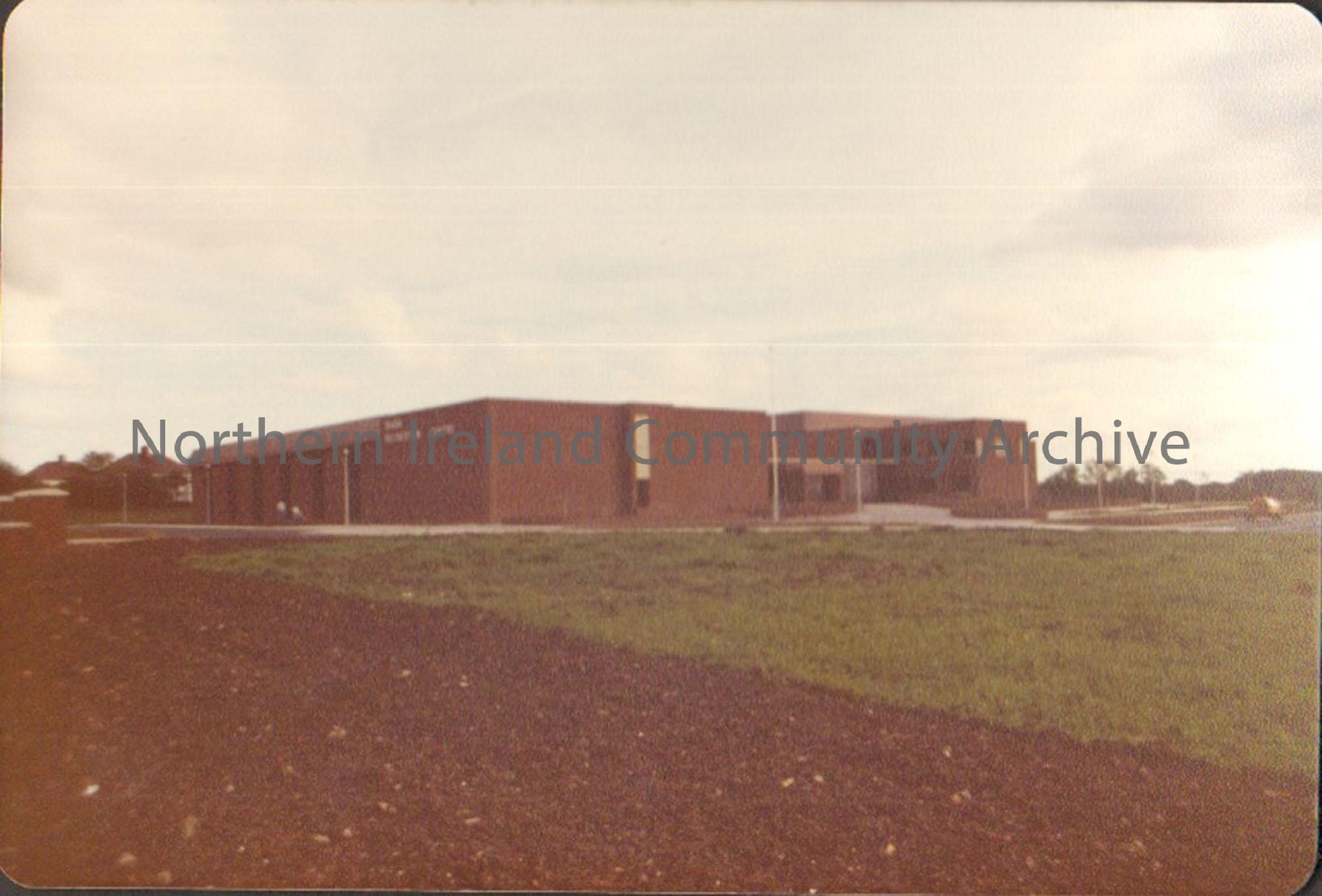 Colour photograph of Riada Recreation Centre. Large brown square building with ploughed land and recently planted grass in front of it.