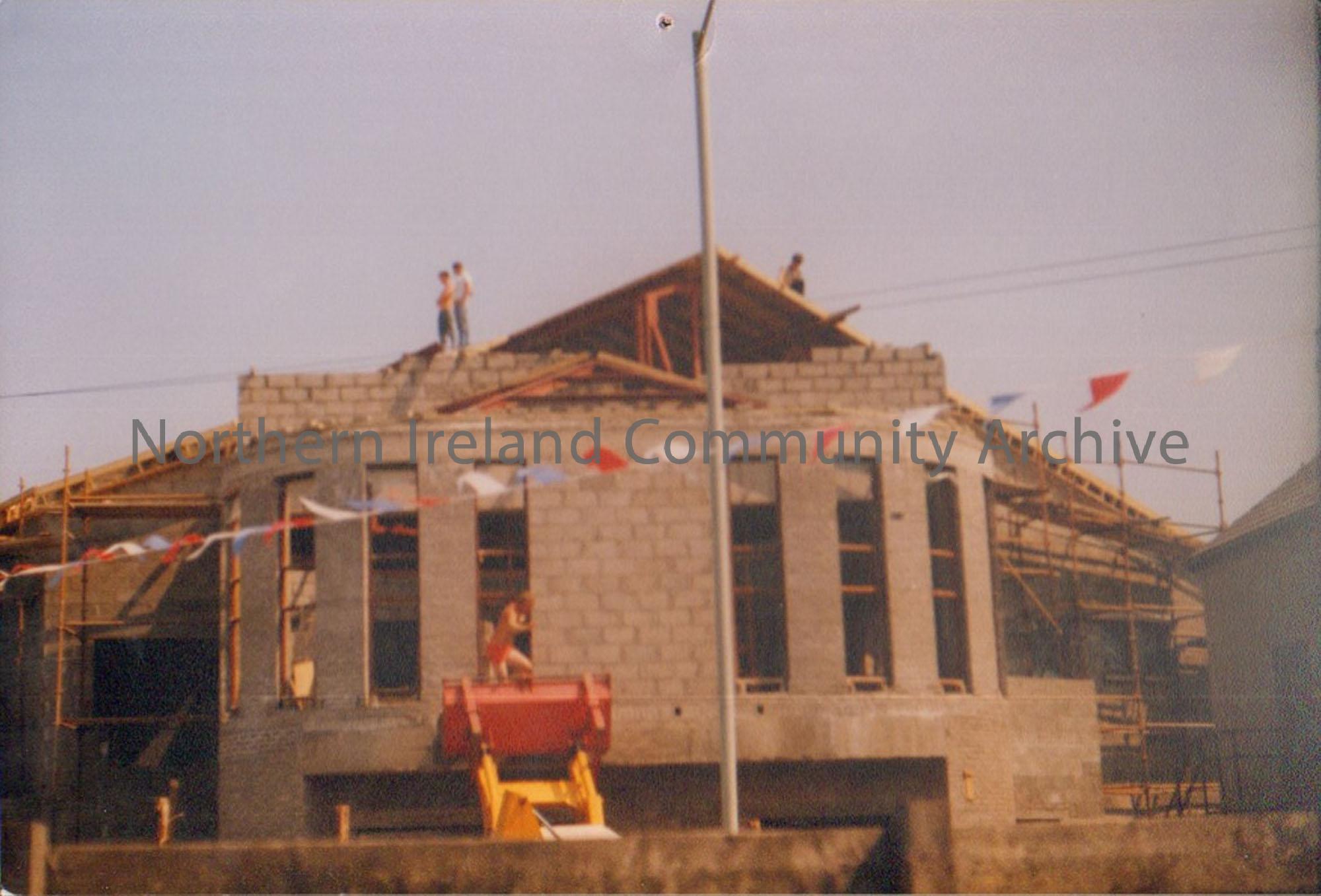 Colour photograph of the building of Riada House Extension. Grey building with no glass in the windows. people standing on top of a part built wall. Y…