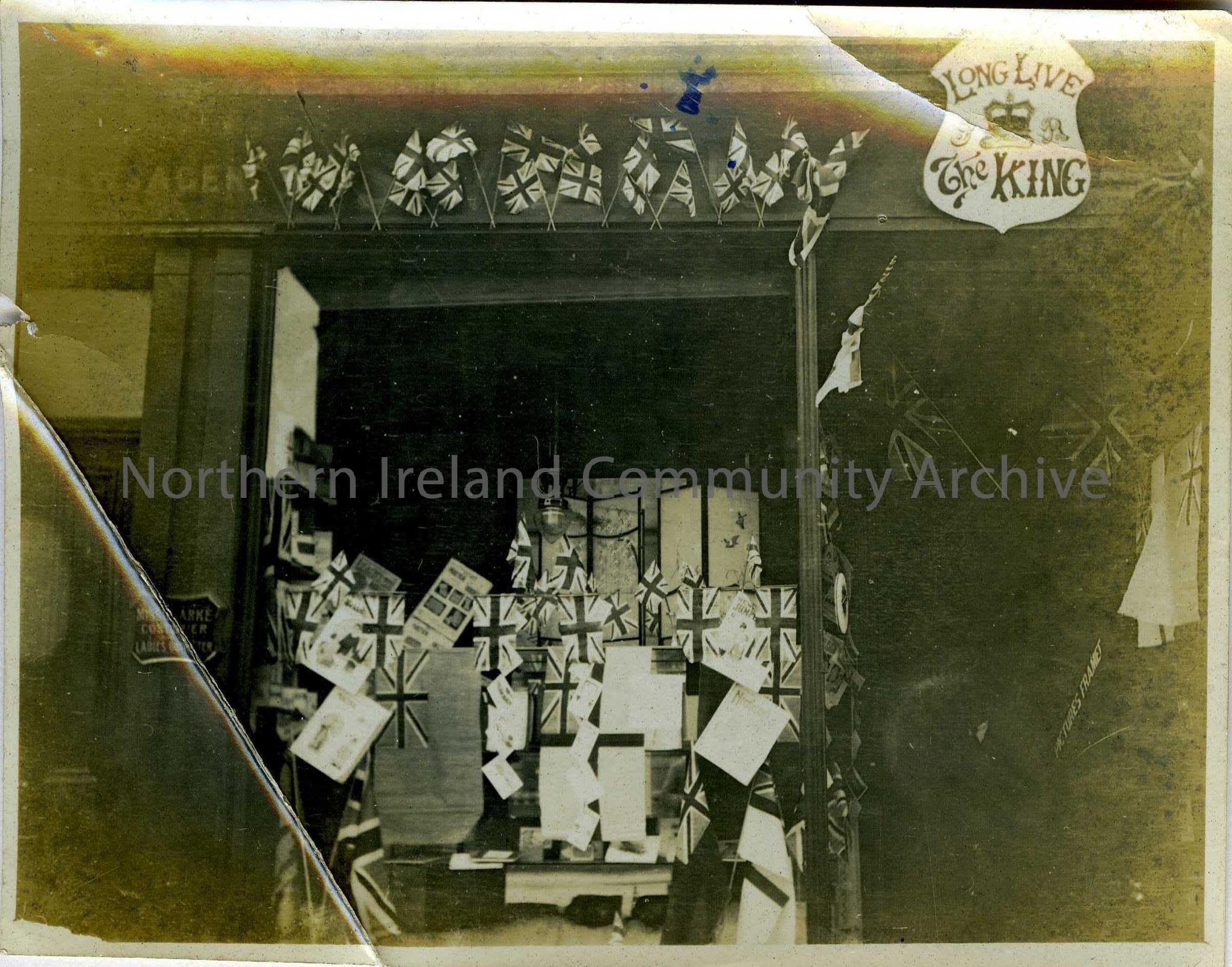 Black and white photograph of a shop front (newsagents?) decorated with union jack flags. Plaque above the door reads – ‘Long Live the King’. Sign on …