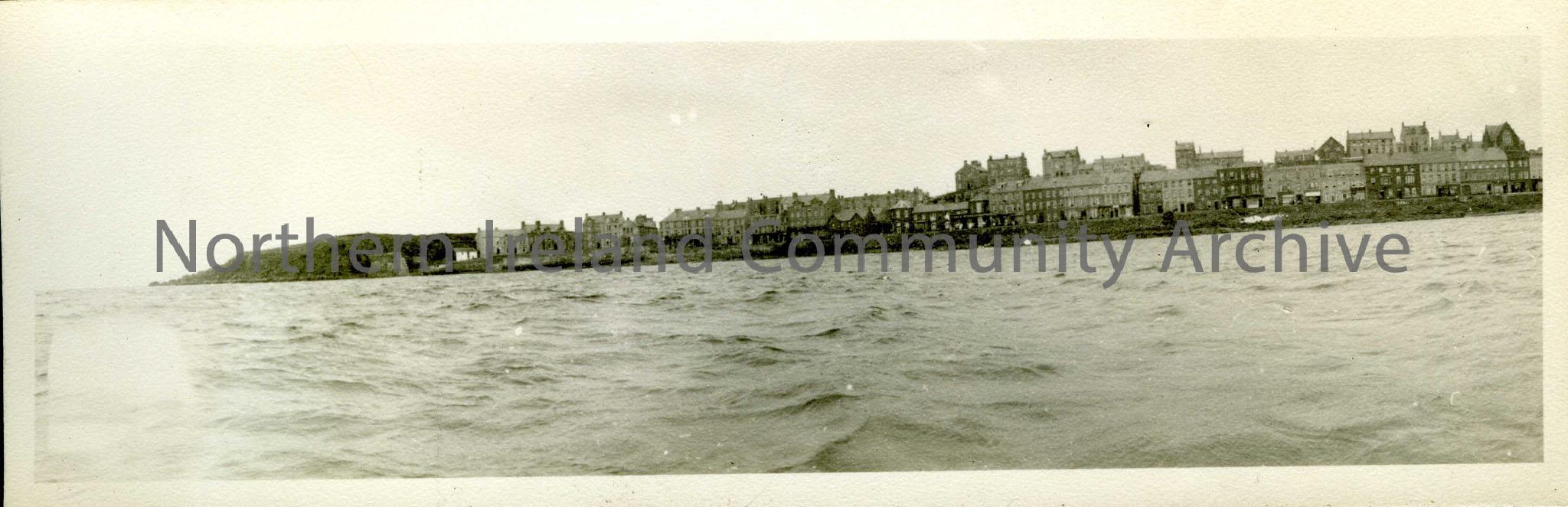 Black and white photograph of Portstewart promenade. Taken from a boat on the sea. The Harbour is visible, with Harbour Hill behind.