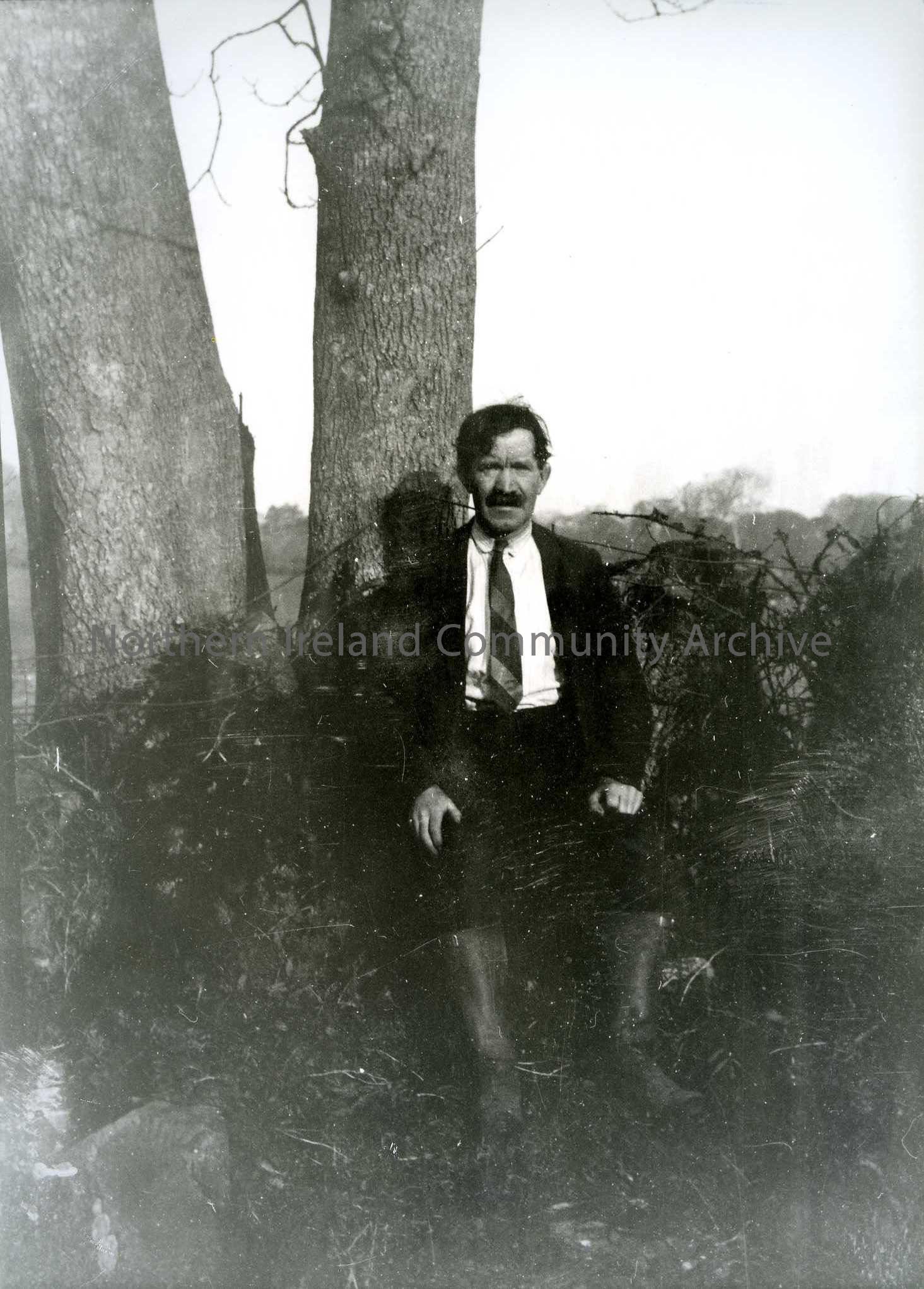 Printed black and white photograph – Man in shirt and tie wearing wellington boots sits in front of trees.