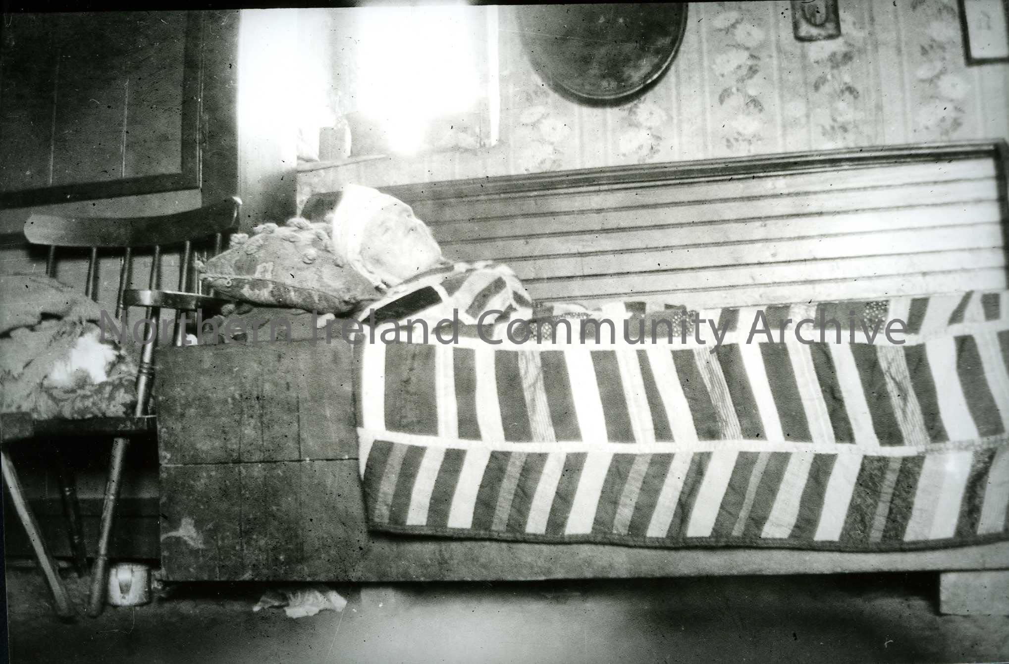 Printed black and white photograph – Elderly man lying on a bench inside a house, covered in a blanket and wearing a hat.