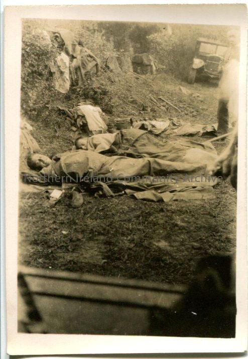 Black and white photograph of men lying under blankets in a ditch. Vehicle in the background.
