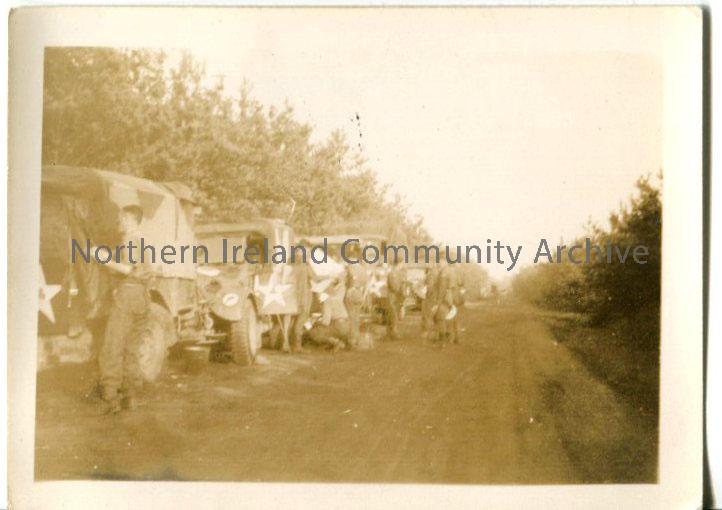 Sepia photograph of army vehicles parked on the side of road. Road has high hedges either side. Group of men next to the vehicles, one is consulting a…