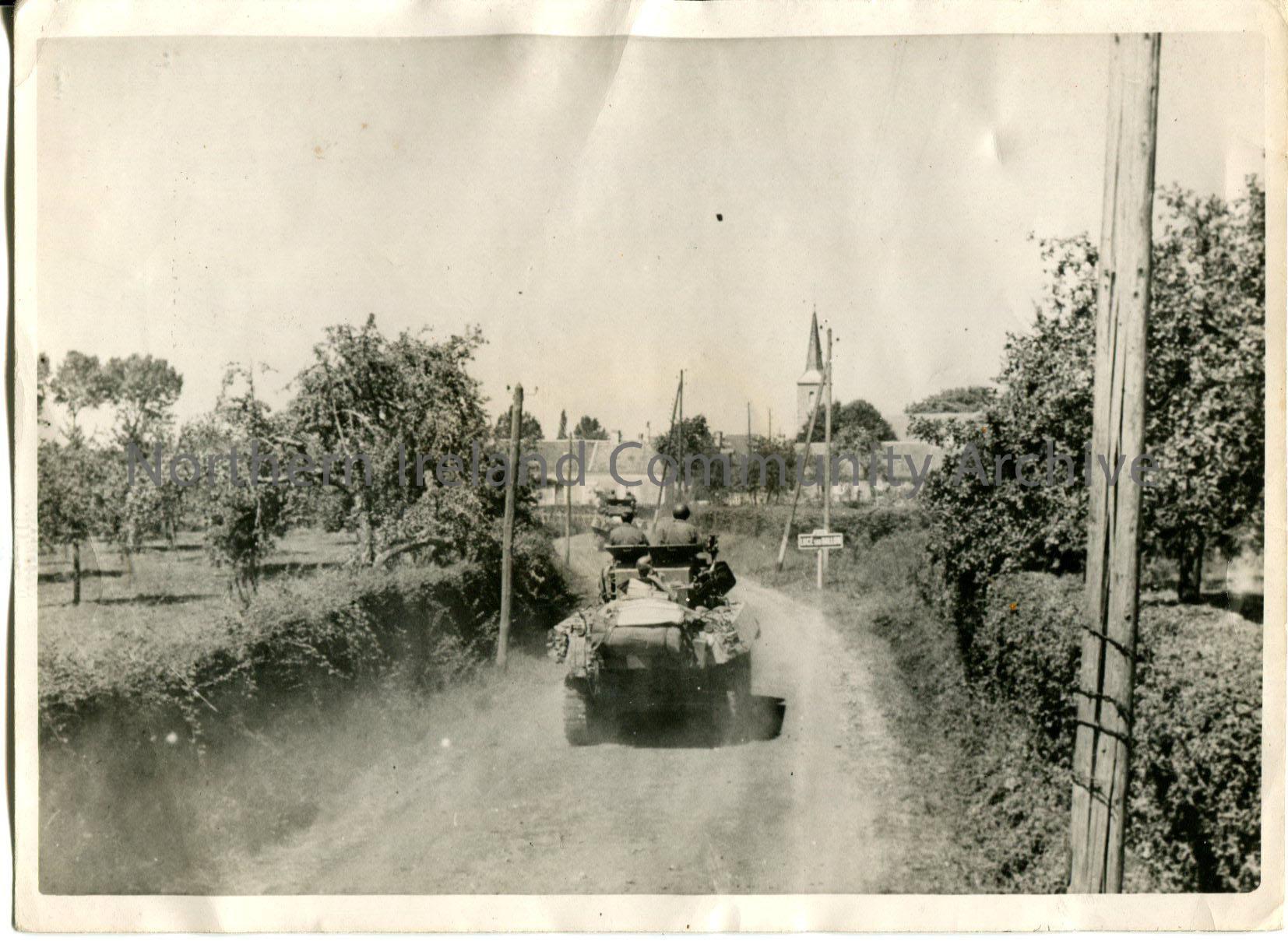 Black and white photograph of a tank with two men on board driving through the countryside towards a village with a church in the distance.