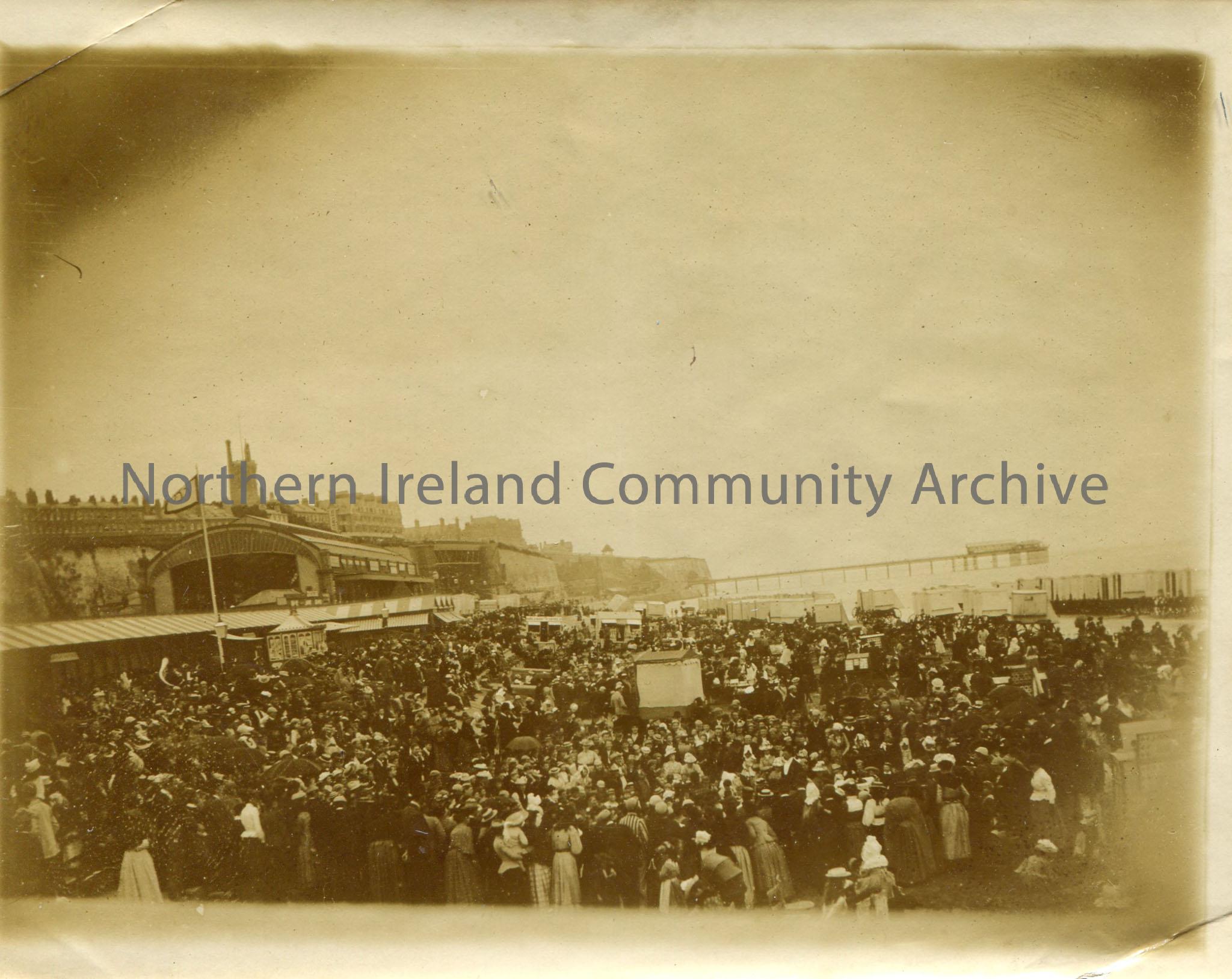 Black and white photograph of a large crowd, possibly on a beach with a pier in the background.