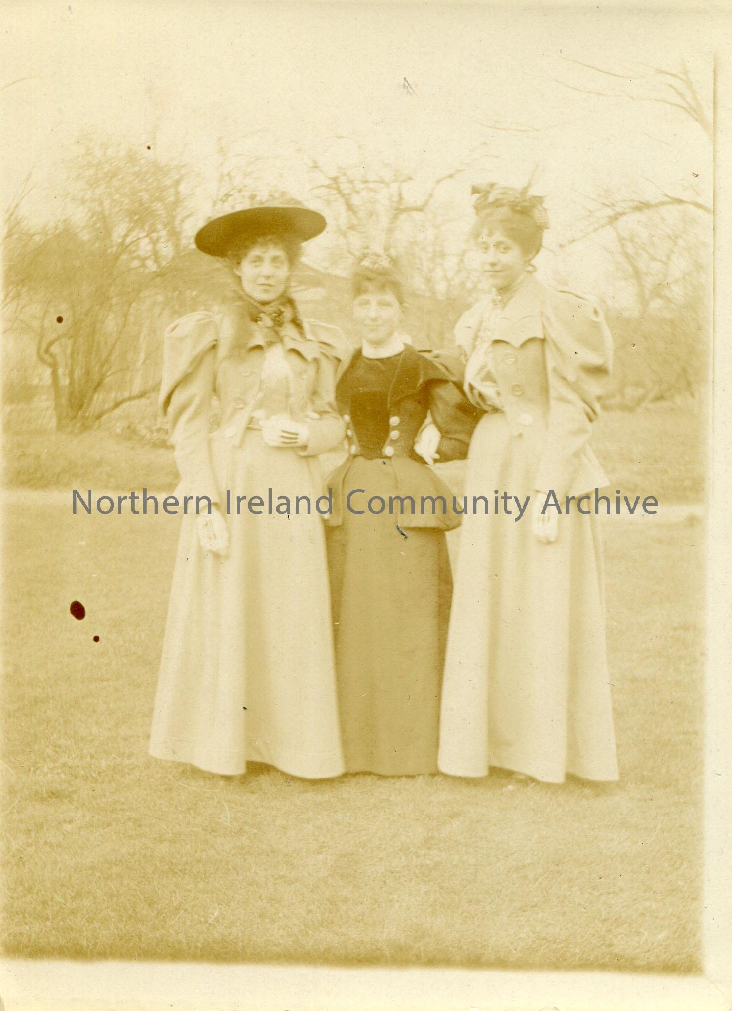 Black and white photograph of three women interlinking arms standing on grass. Trees with no leaves in background.