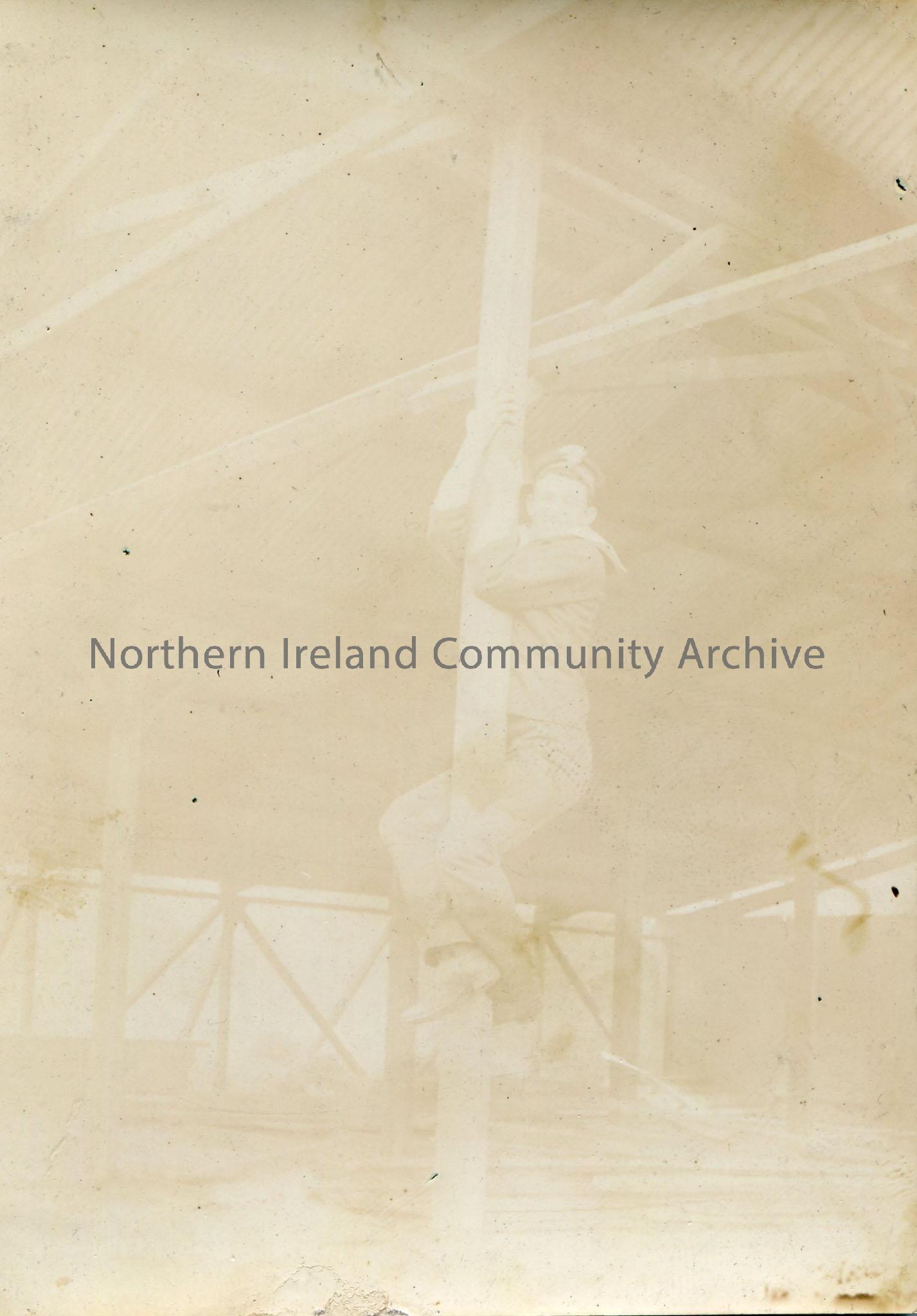 Black and white photograph of a young man climbing a wooden pole, possibly wearing a sailor or Navy uniform. Image very faded.