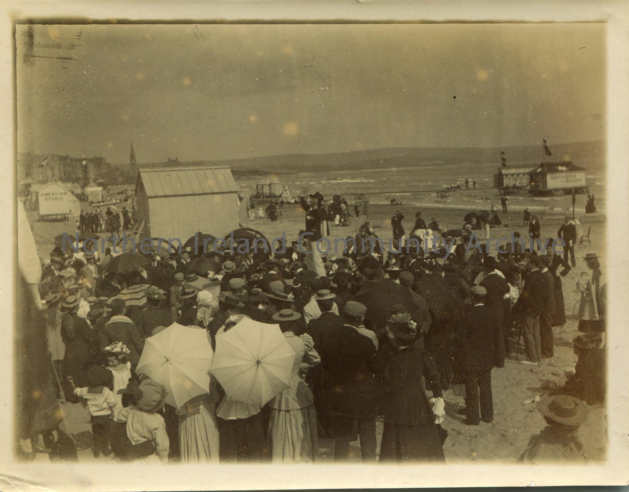 Black and white landscape photograph of a crowd at a beach watching a man perform. Gentleman’s Saloon hut in the sea in the background.