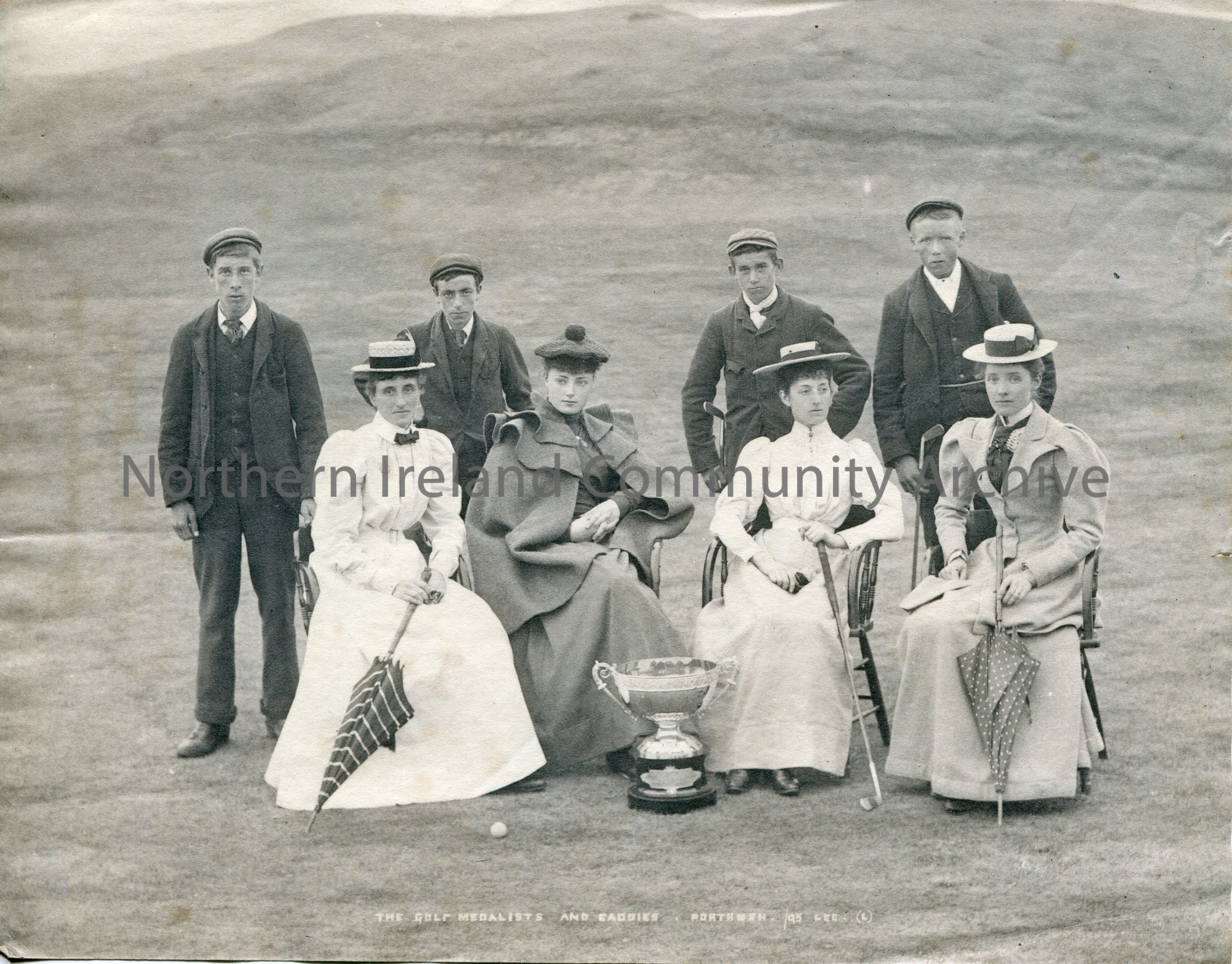 Black and white photograph of 4 women golfers sitting on chairs on a golf course in all their finery in front of their caddies who are in white collar…