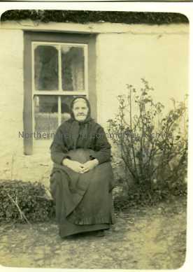 Photograph of an elderly lady sitting outside a cottage.
