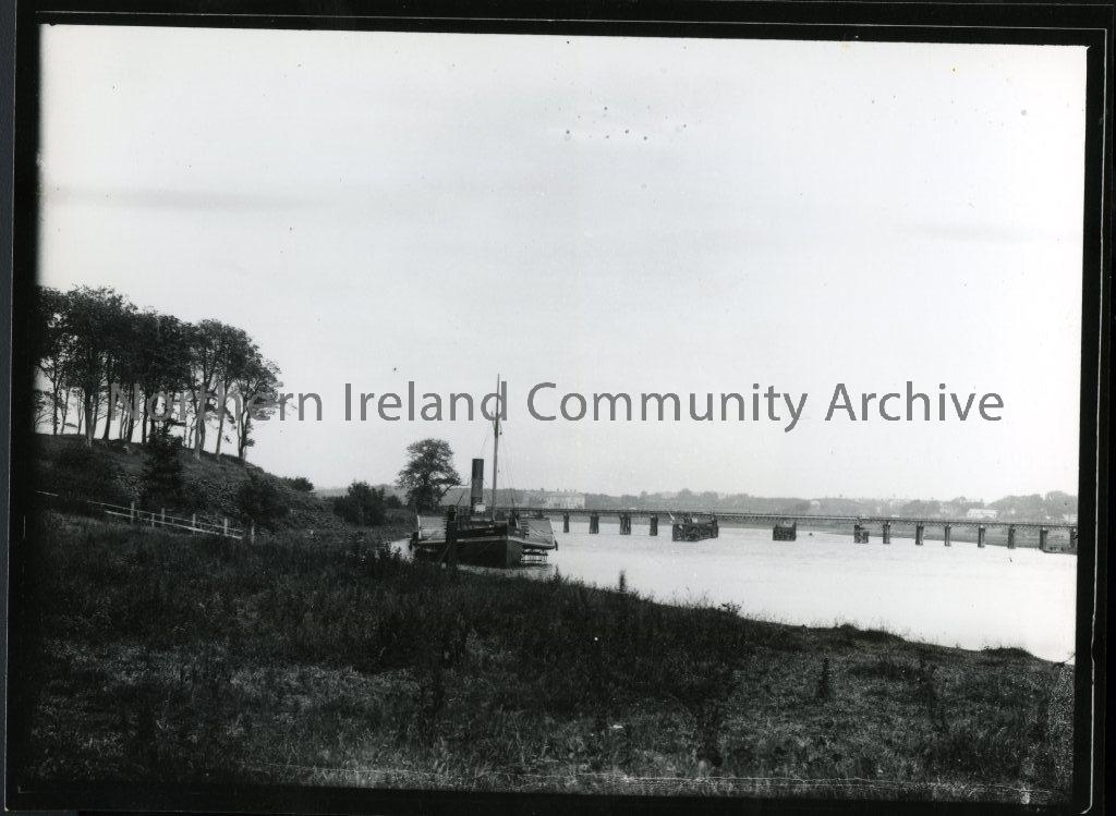 Black and white photograph view of the River Bann and train bridge, Coleraine. Paddle steamer Confidence beside County Hall Grounds with old railway bridge in the background.  (5687)