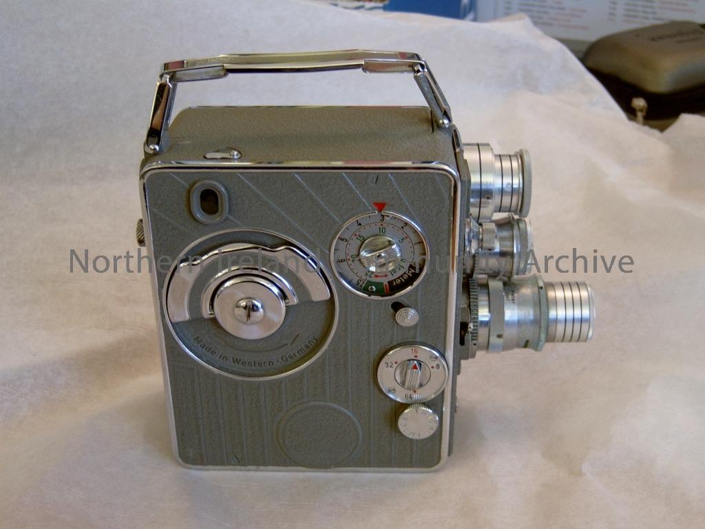 Grey super 8 cini camera with 3 protruding dials at the front including lens and focus. Make is Nizo Heliomatic 8 Mod.S2R. On one side of the camera is the label of the make with technical details. On the other side are the dials for using the It is label (5055)