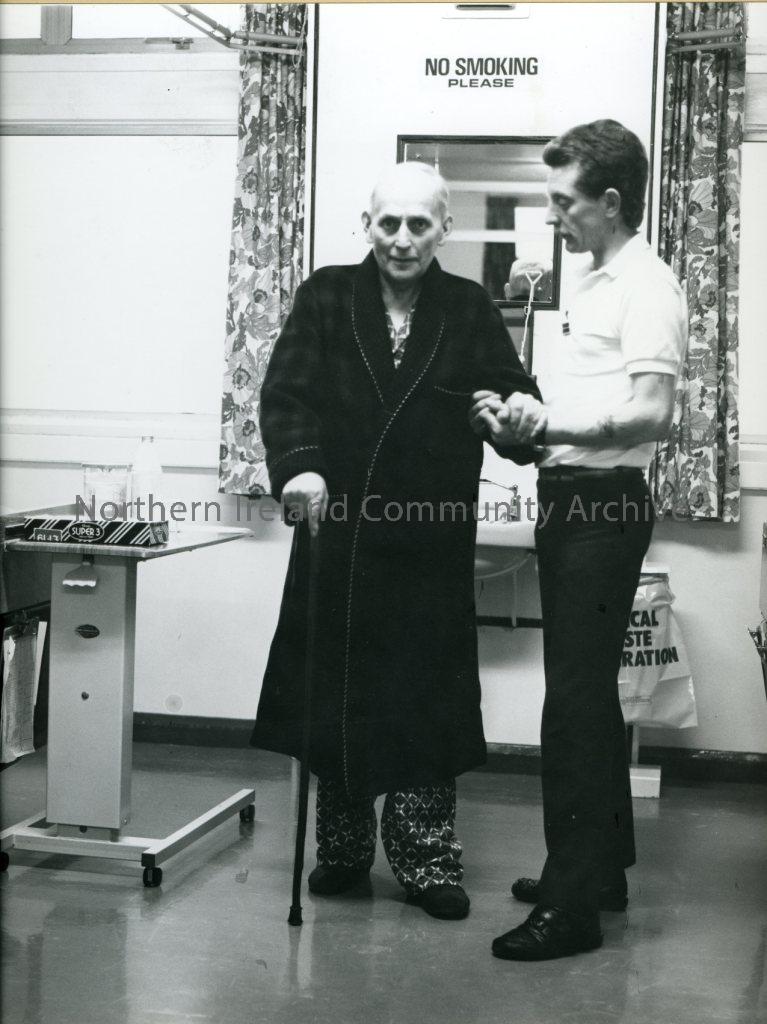 black and white photograph of a man aiding an elderly man with a walking stick in a hospital (3306)